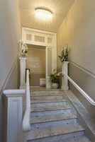 Una scalinata in marmo con ringhiere bianche e un lampadario, che conduce a una porta con una targa che riporta la scritta 'Welcome to Villa Aria'.