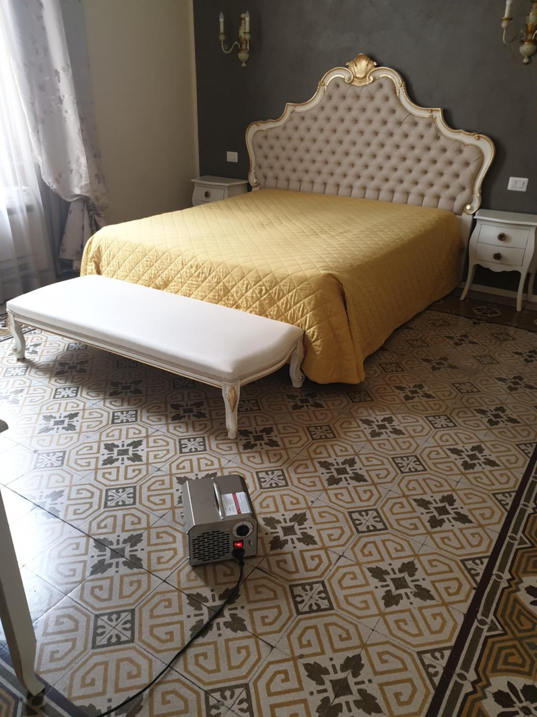 A bedroom with a large bed, a bench, patterned floor tiles, and a heater on the floor.