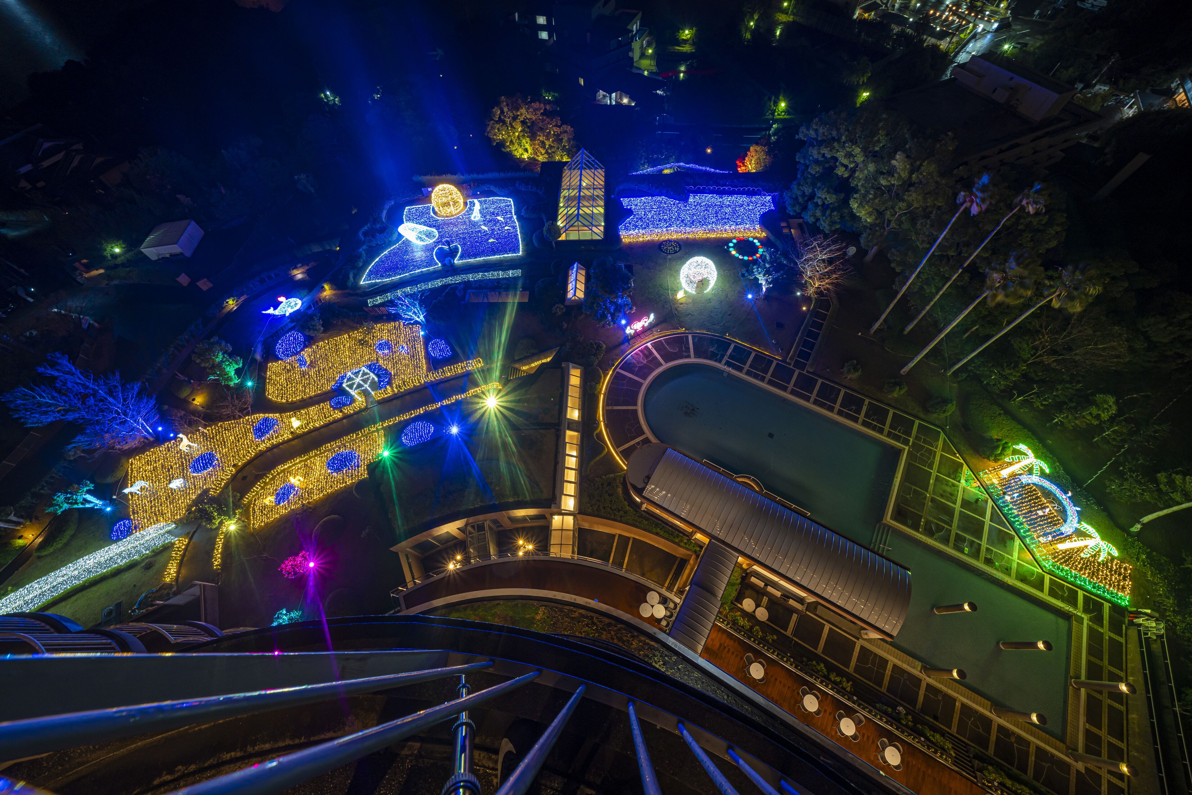 Vista nocturna de un edificio con senderos e instalaciones iluminadas, incluyendo una escalera en espiral y una zona de piscina.