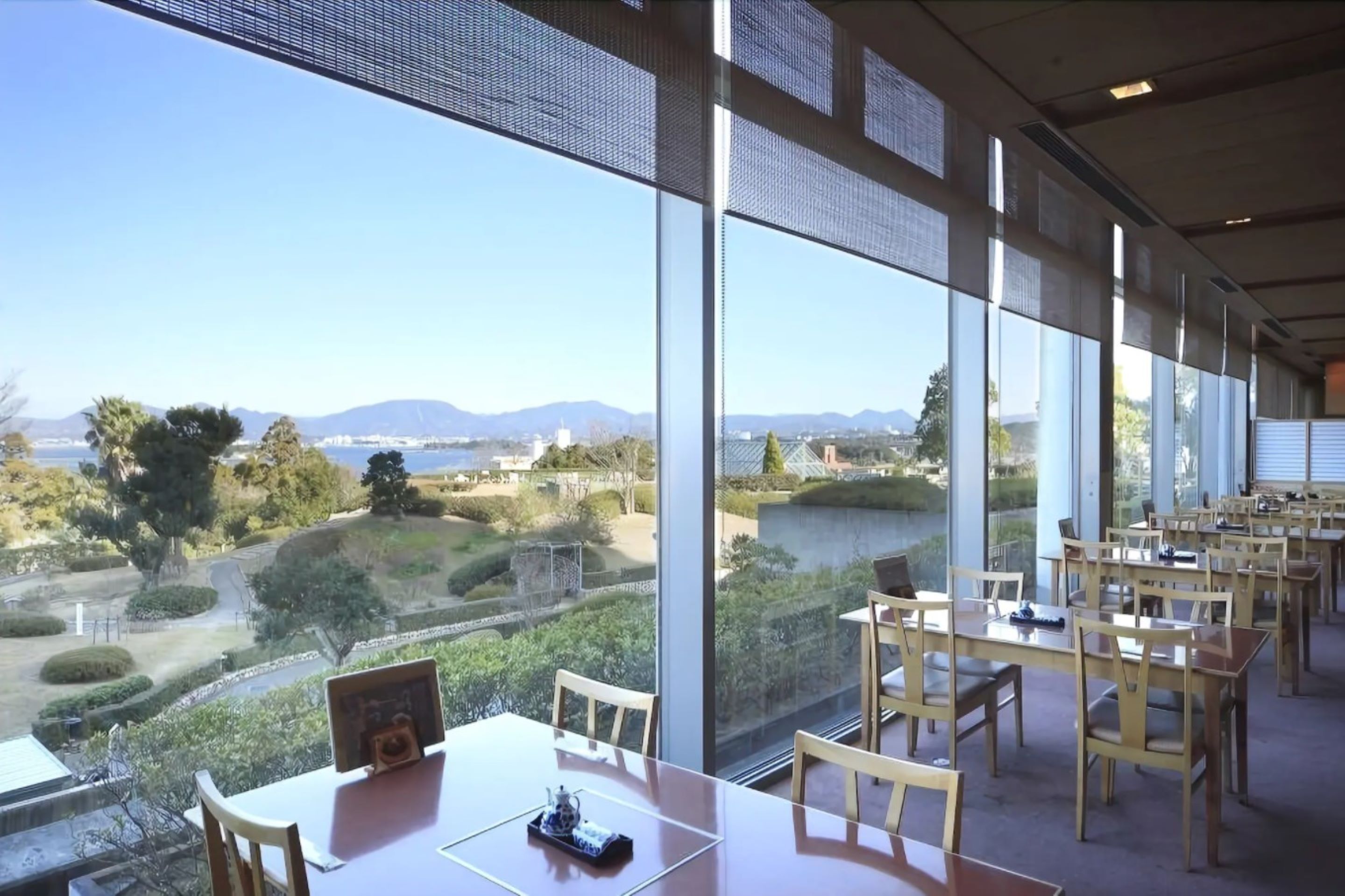 Un comedor con mesas y sillas, amplias ventanas que ofrecen vistas a un jardín ajardinado y montañas lejanas.