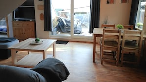 Ein gemütliches Wohnzimmer mit einer Couch, einem Esstisch mit Stühlen, einem Fernseher auf einem Holzschrank und einer großen Schiebeglastür, die zu einer überdachten Außenbereich führt.