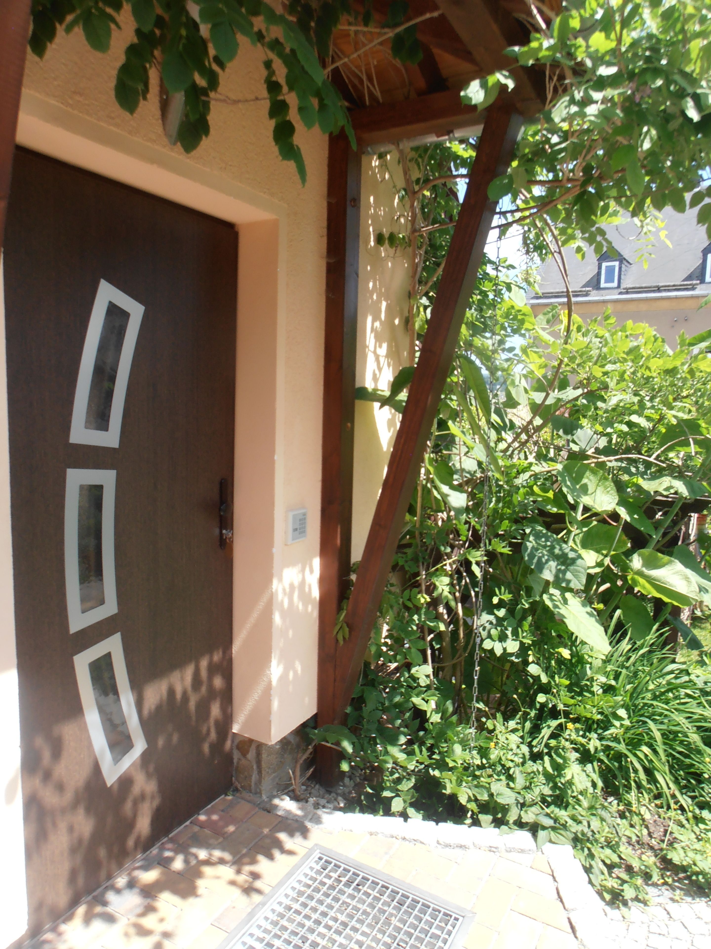 Una puerta de madera con tres abertura rectangulares, una rejilla de metal y un jardín con varias plantas y un árbol.