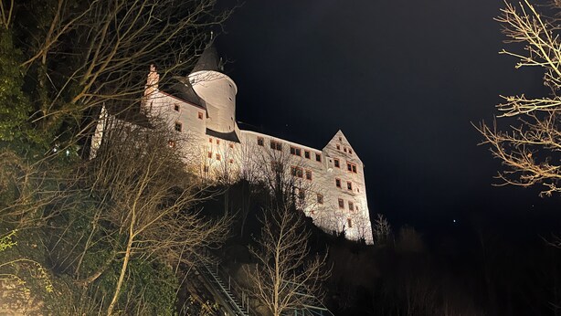 Ein großes, schlossartiges Gebäude mit zahlreichen Fenstern und einem markanten Turm, umgeben von Bäumen und einer Treppe, die dorthin führt.