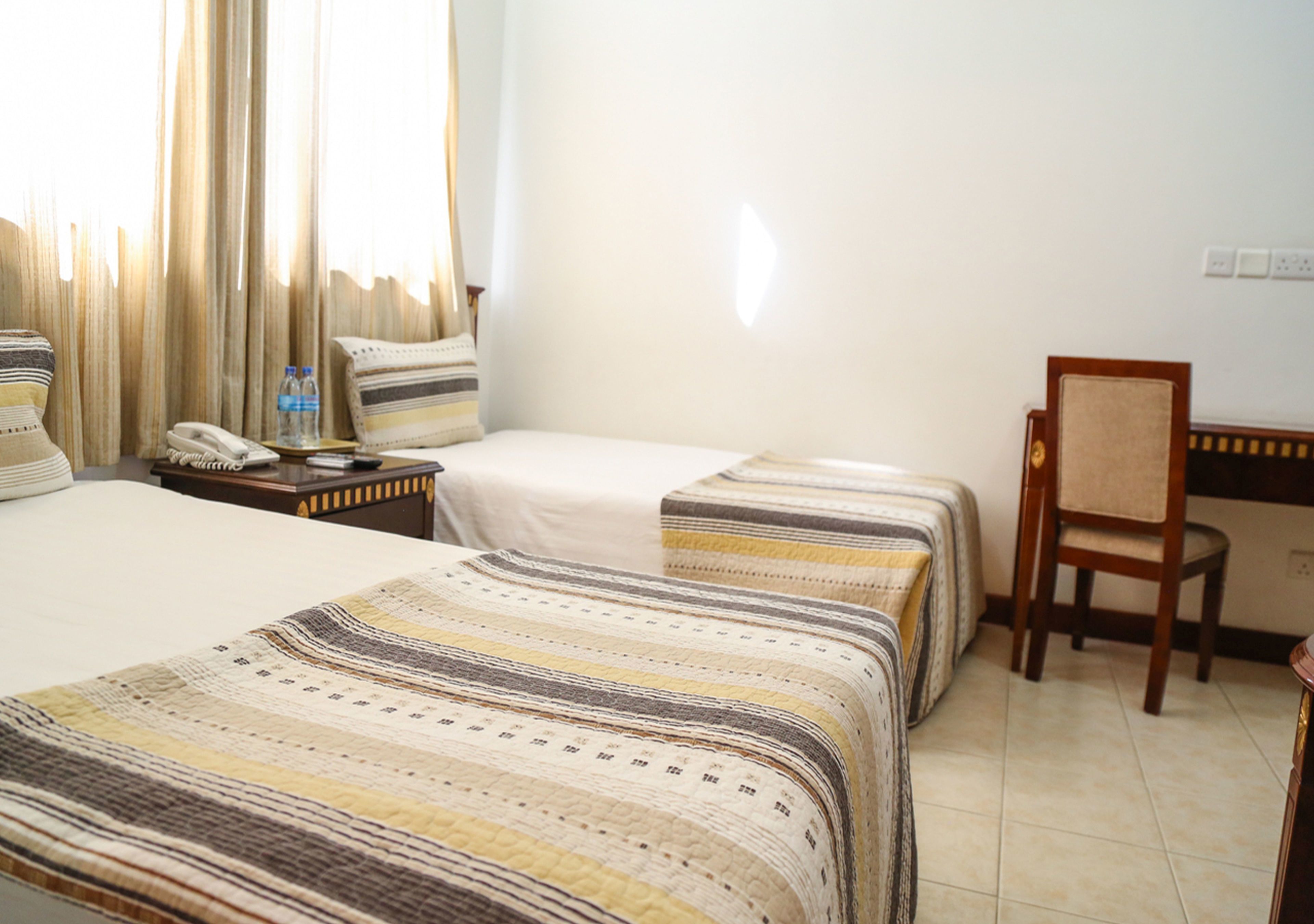 Une chambre d’hôtel avec deux lits, une chaise, une table de chevet avec un téléphone et une fenêtre avec des rideaux.