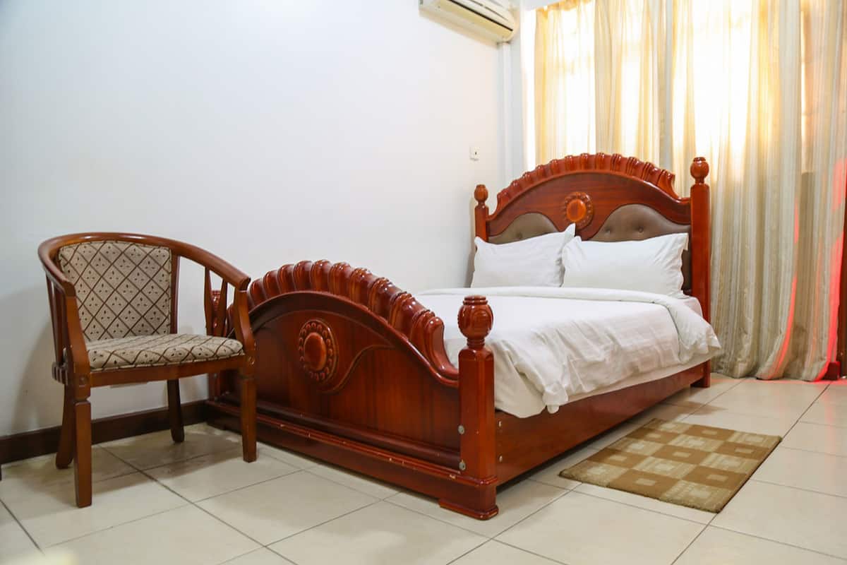 Un lit en bois avec une tête de lit, une chaise en bois avec un siège rembourré et un tapis à motifs posé sur un sol carrelé.