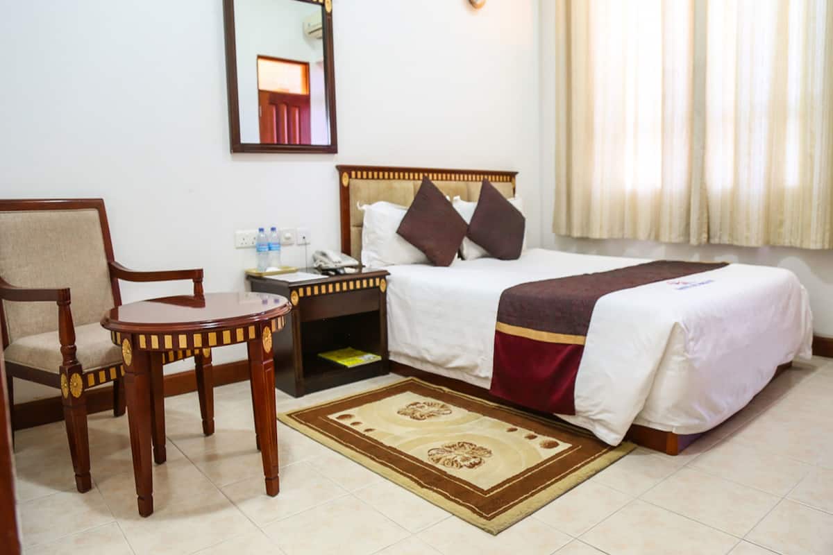 Une chambre d’hôtel avec un lit, une chaise, une table de chevet, un miroir et une fenêtre avec des rideaux.