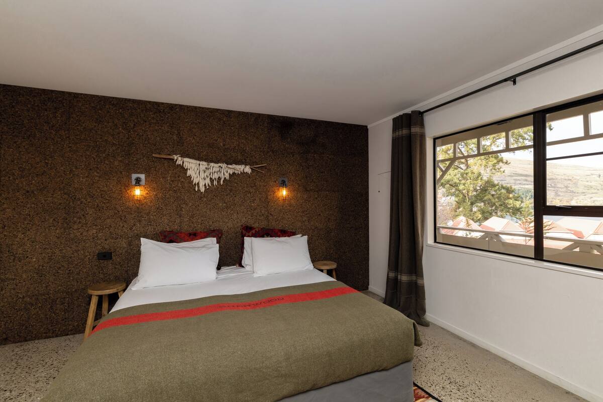 A bedroom with a large bed, two bedside tables, a wall-mounted light fixture, and a window with curtains.