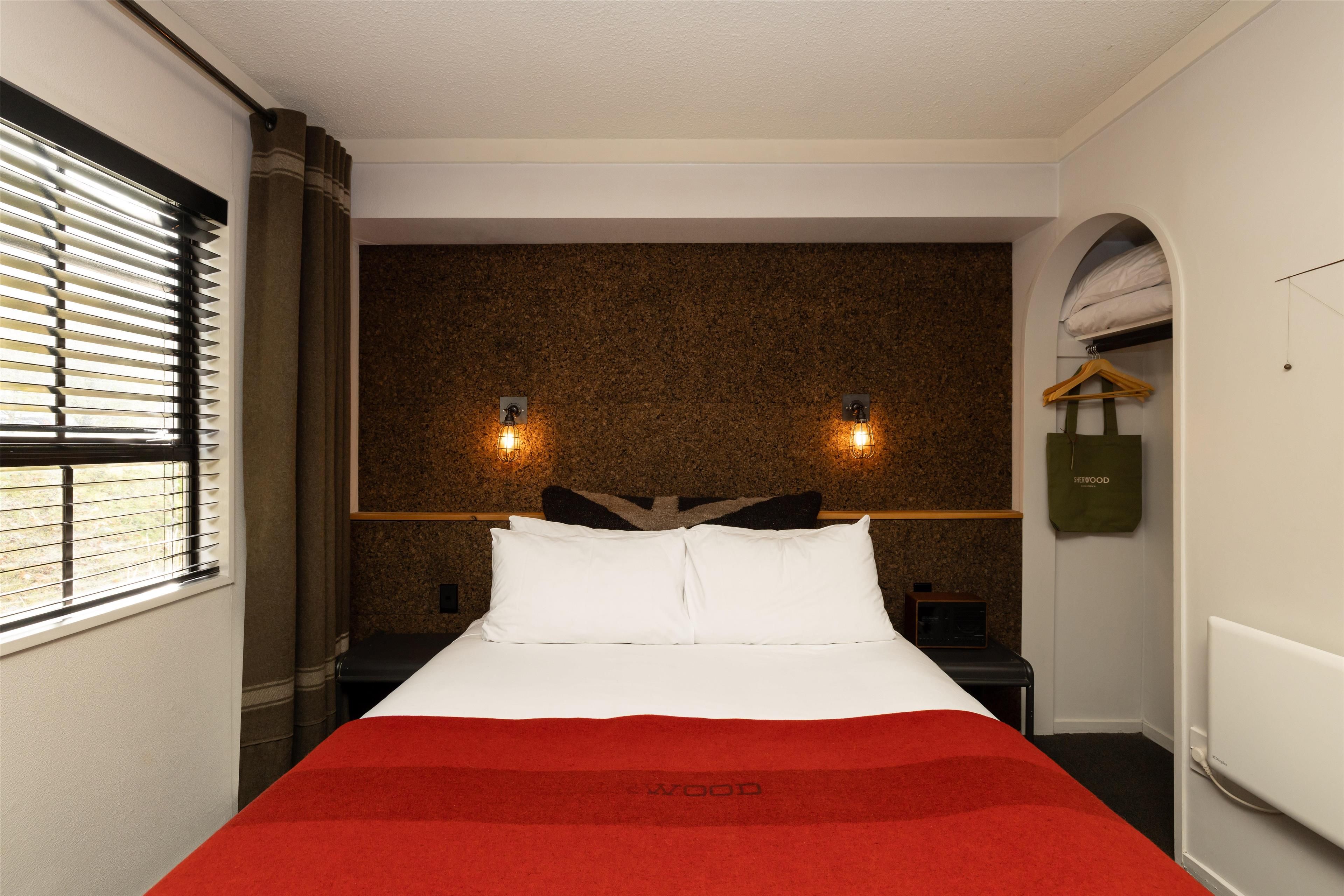 Una habitación de hotel con cama, mesita de noche, lámpara de pared y ventana con persianas.