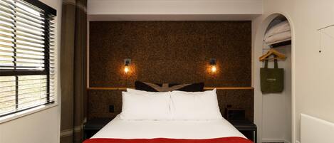 Una habitación de hotel con cama, mesita de noche, lámpara de pared y ventana con persianas.
