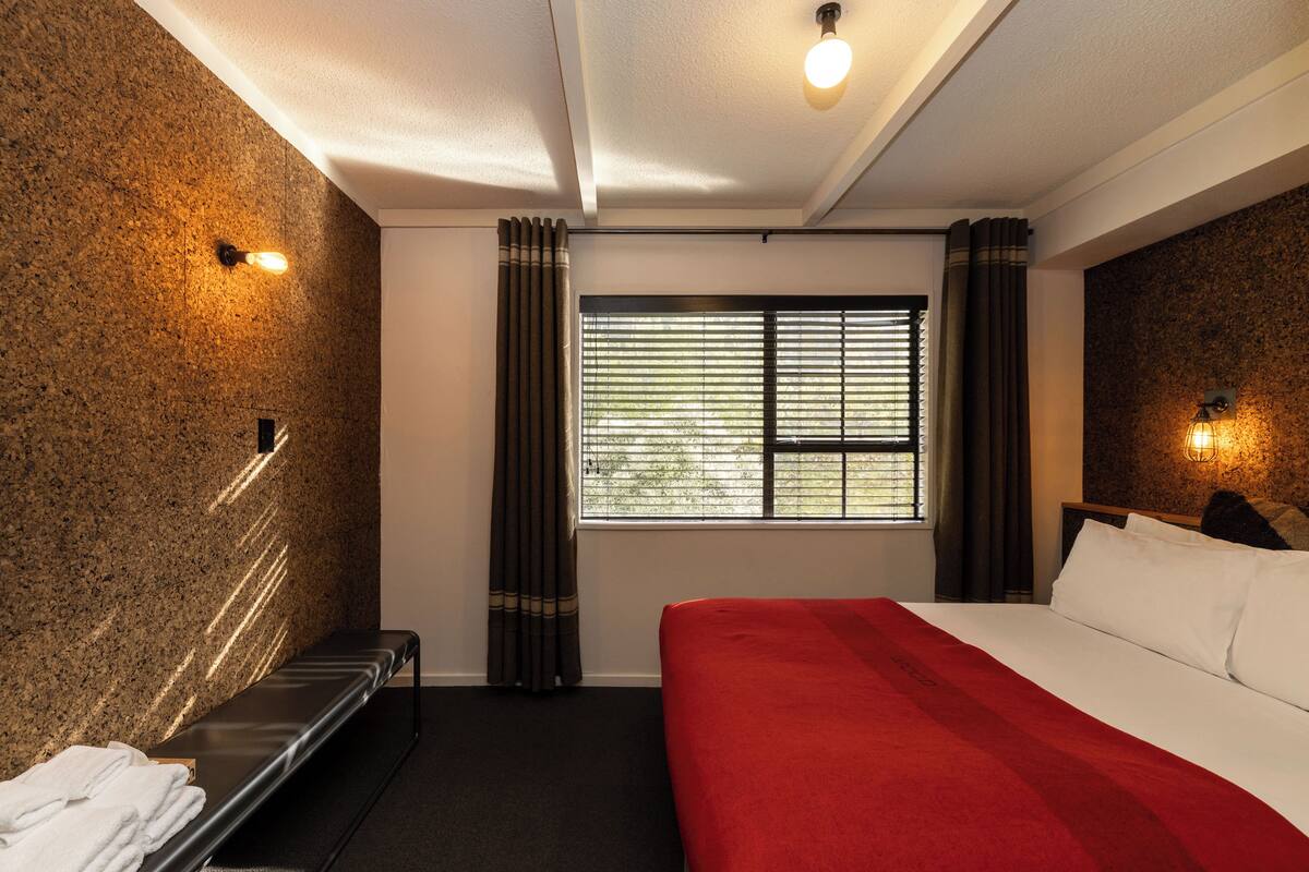A hotel room with a bed, a bench, a window with curtains, and wall-mounted lights.