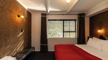 Quarto de hotel com uma cama, um banco, janela com cortinas e luminárias fixadas na parede.