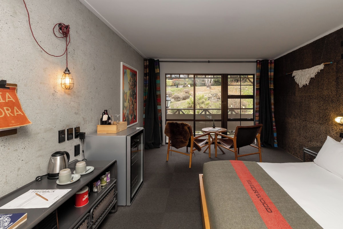 A hotel room with a bed, a desk with a kettle, cups, and a mug, a chair, a small table, and a window with a view of the outdoors.