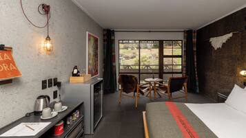 Habitación de hotel con cama, escritorio con hervidor, tazas y una mug, una silla, una mesita y ventana con vistas al exterior.