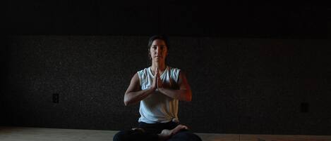 A person sitting cross-legged on a mat in a dimly lit room with a plain wall and a wooden floor.