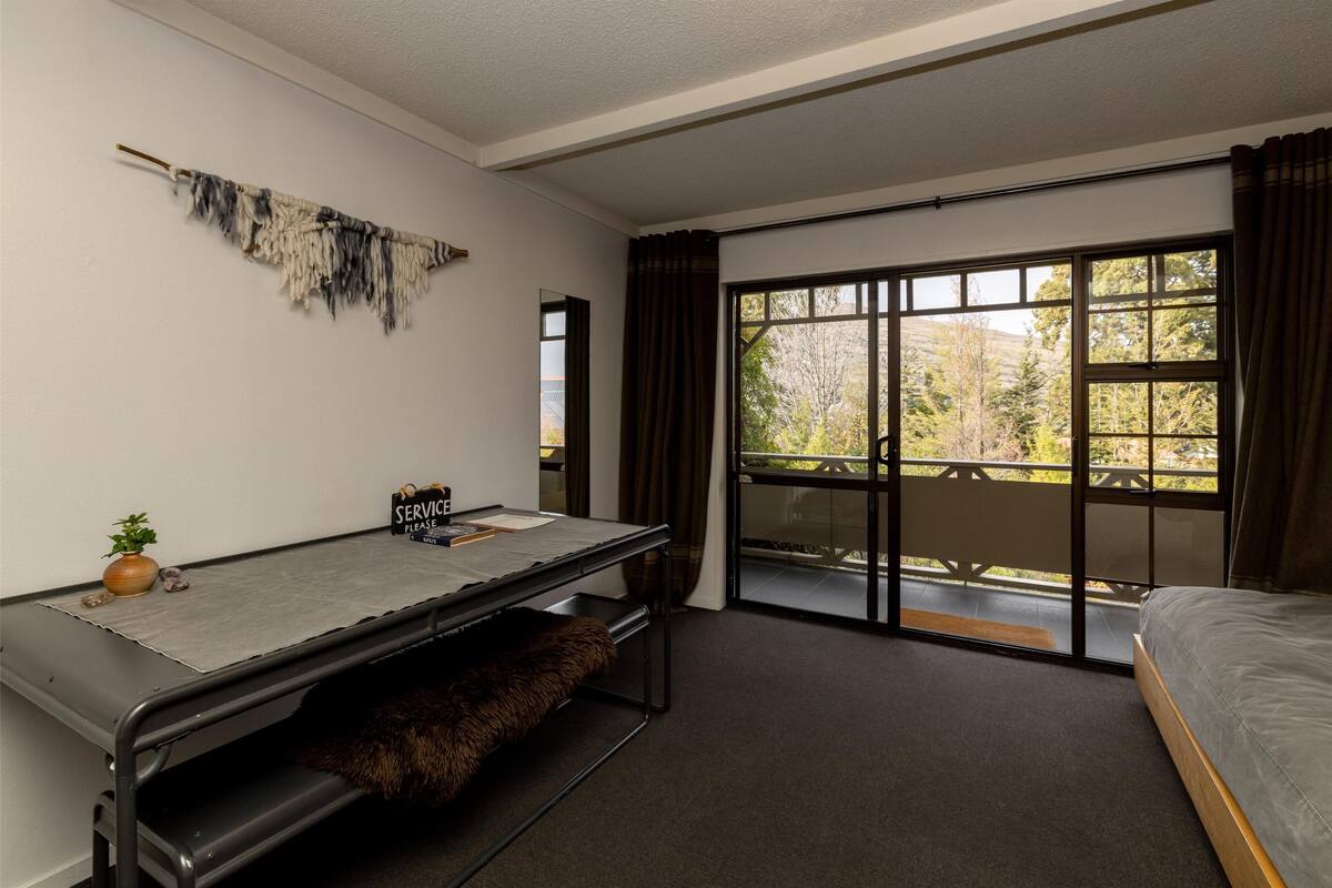 A bedroom with a bed, a desk with a chair, a large window with curtains, and a view of trees outside.