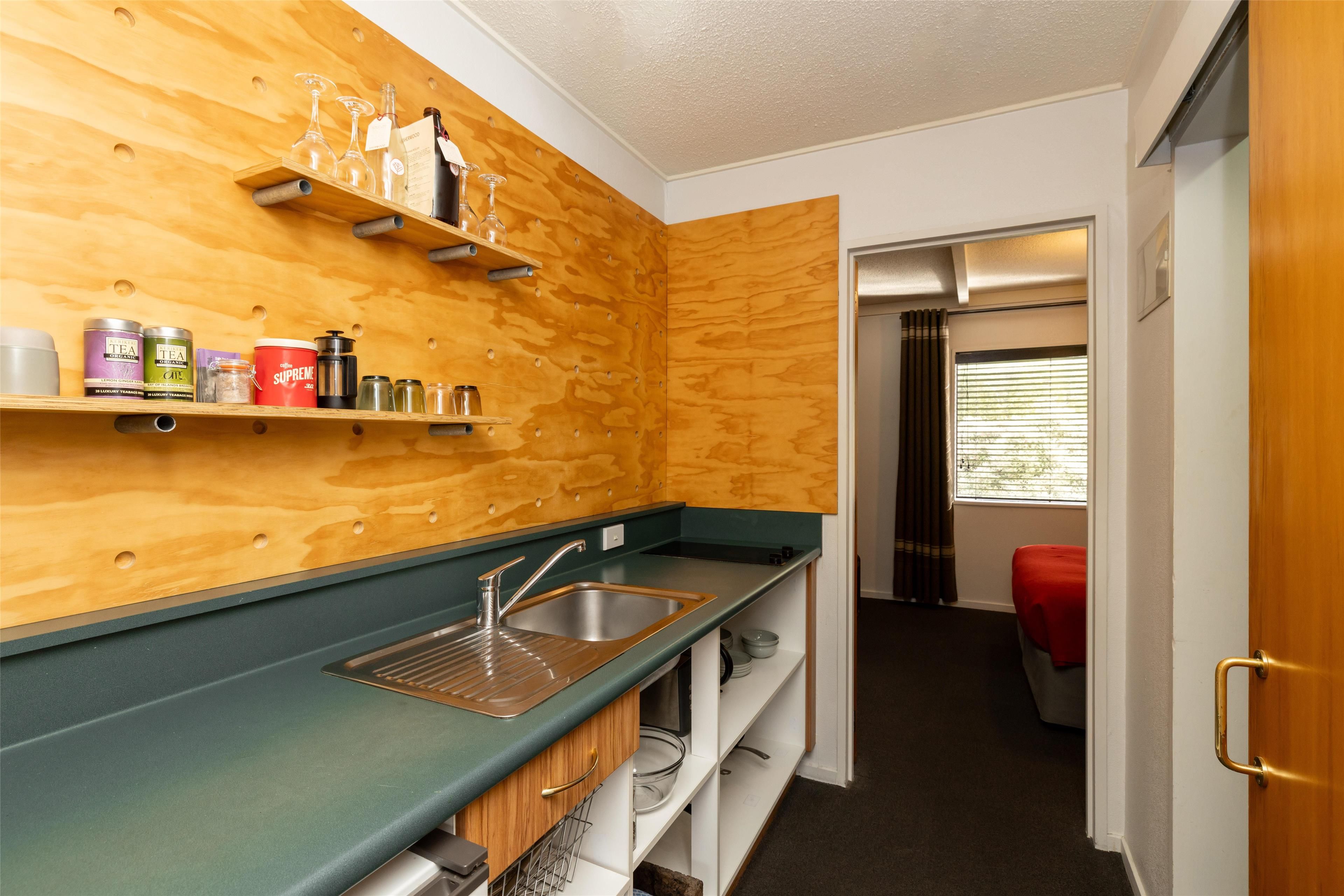 Una cocina compacta con paneles de madera en la pared, una encimera verde y un lavamanos de acero inoxidable. Se observan estantes con diversos recipientes y una pequeña ventana con persianas.