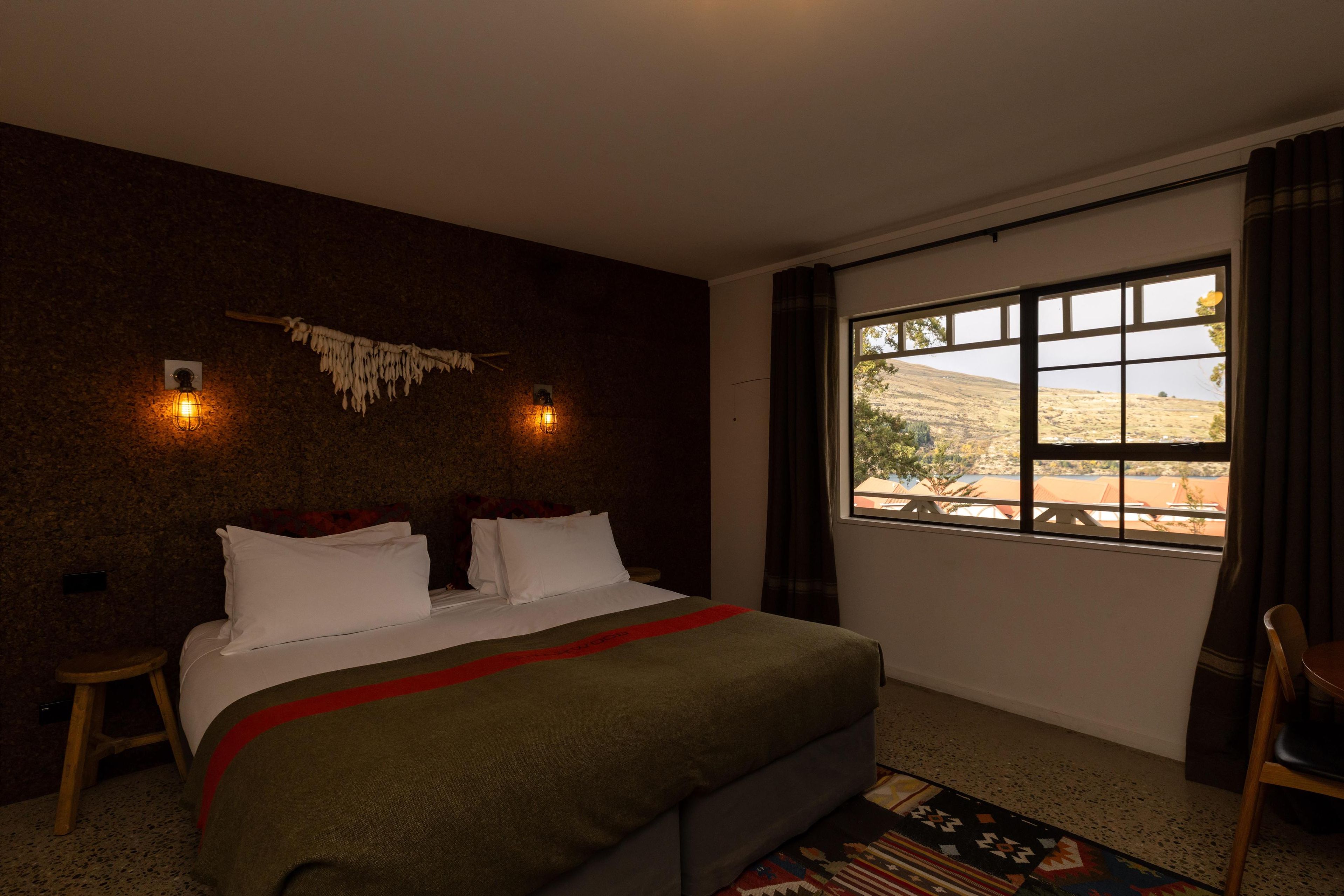 A bedroom with a bed, a window with a view of a landscape, a wall-mounted light fixture, and a small stool.
