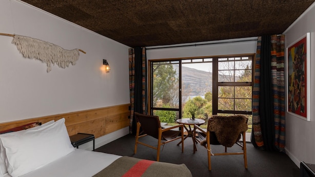 A room with a bed, a wooden headboard, a small table with chairs, a view of the outdoors through large windows, and a wall-mounted light fixture. - Sherwood Queenstown (Queenstown)