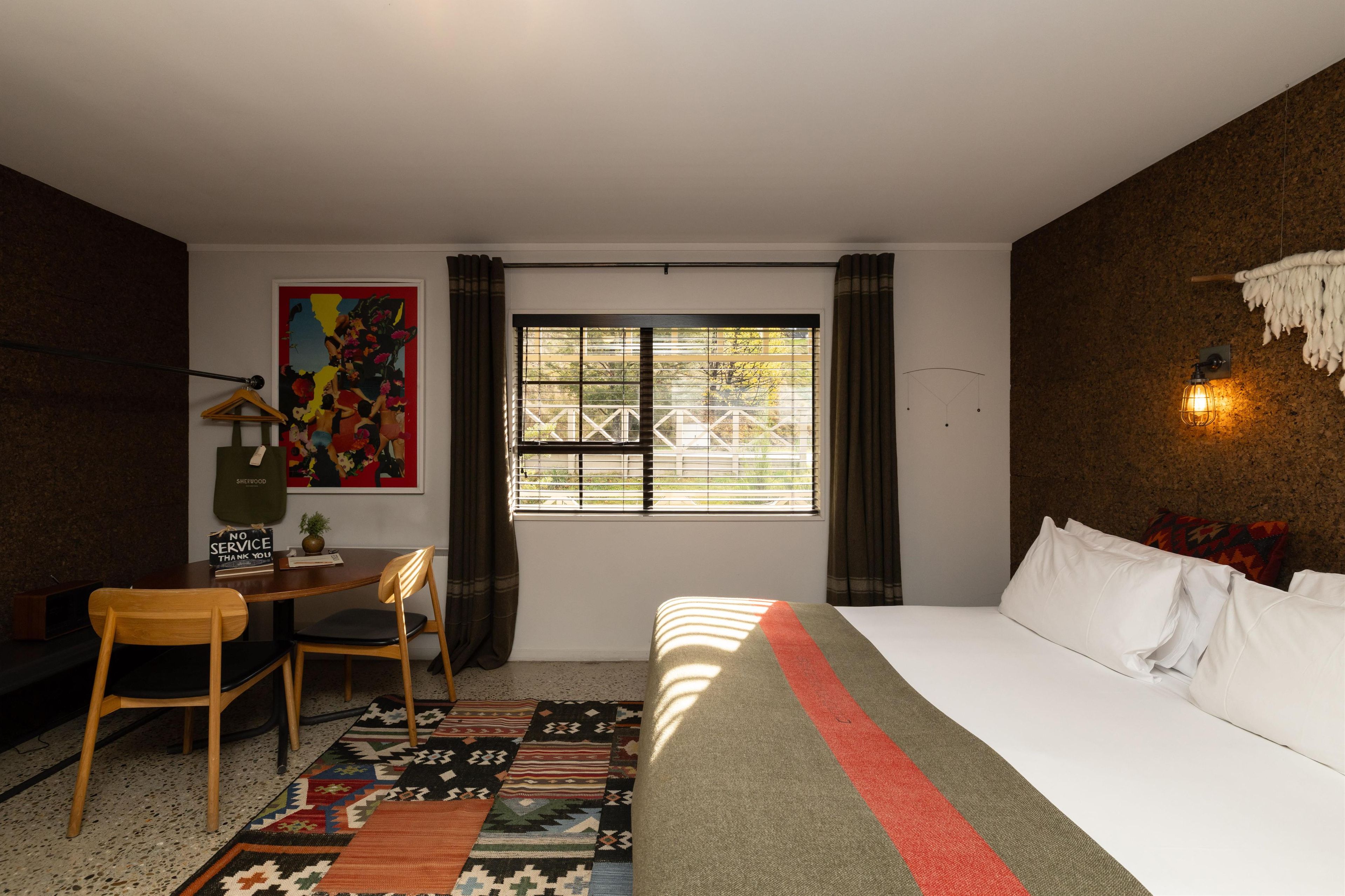 A hotel room with a bed, a desk with a chair, a window with curtains, a colorful painting, and a wall-mounted light fixture.