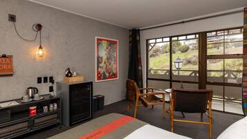 A modern hotel room with a large window, a desk with a kettle, a chair, and a bed.