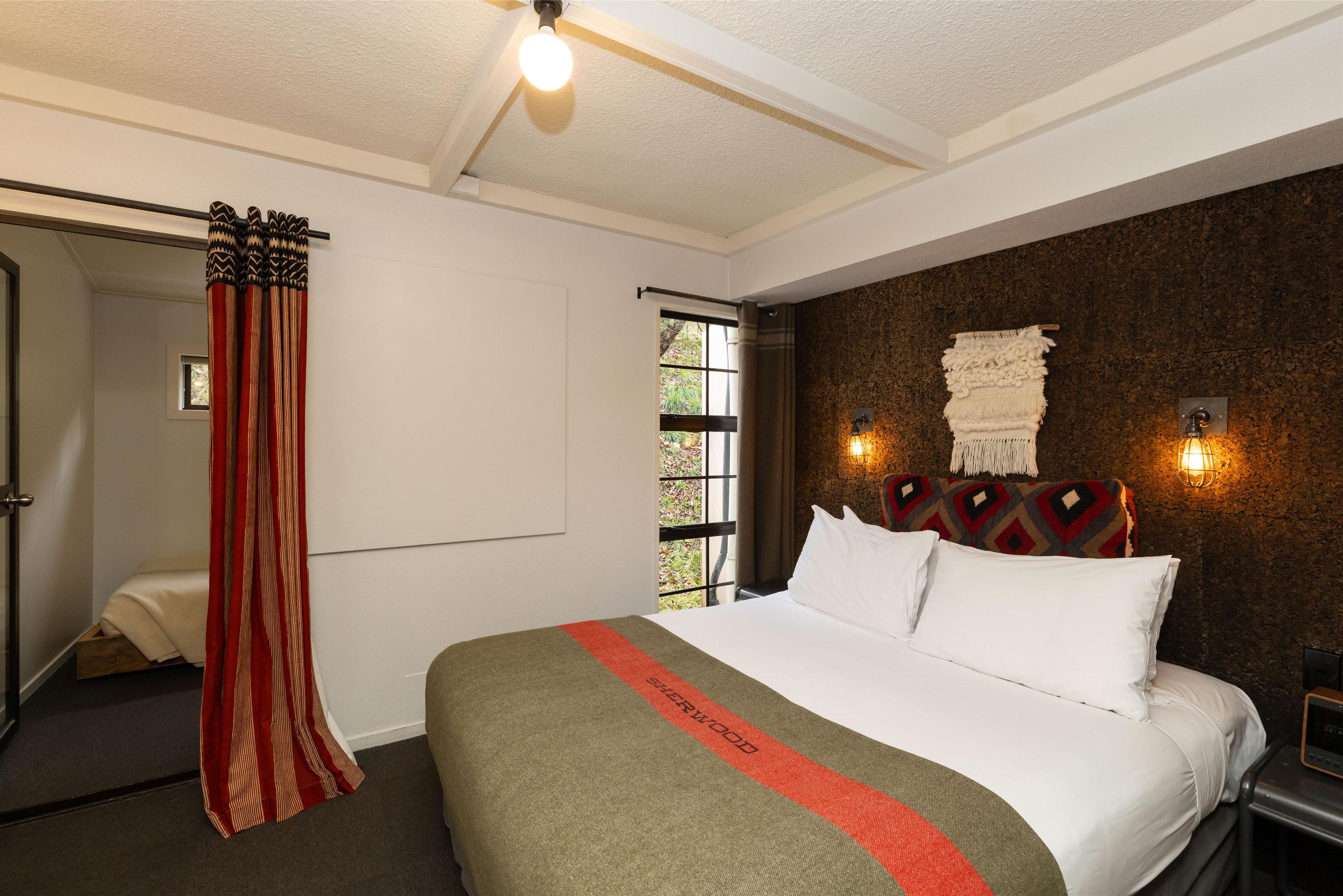 A hotel room with a large bed, a window with a view, a curtain, and a wall-mounted light fixture.