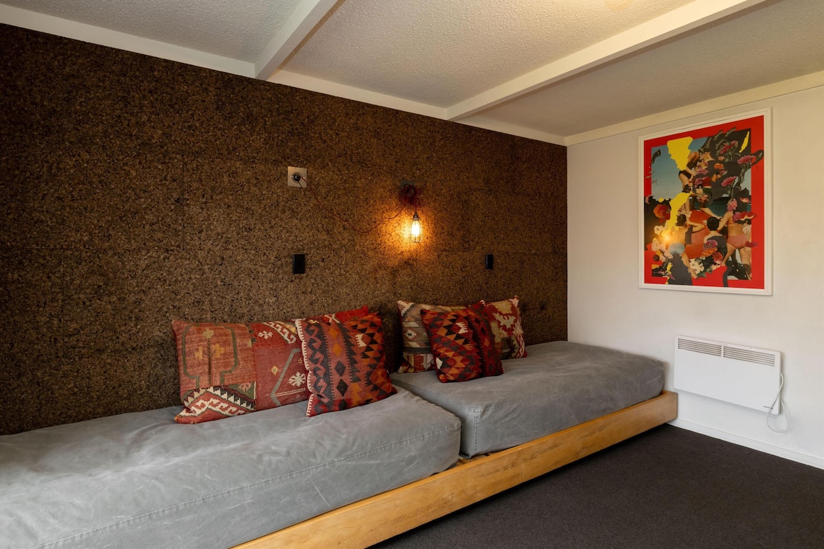 A room with a brown textured wall, a grey sofa with patterned cushions, a wooden platform, and a red and yellow artwork on the wall.