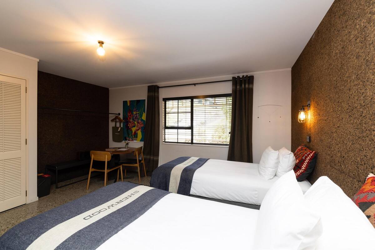 A hotel room with two beds, a desk, a chair, a window with curtains, and a wall-mounted light fixture.