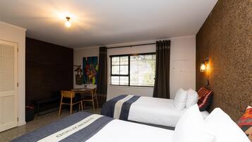 A hotel room with two beds, a desk, a chair, a window with curtains, and a wall-mounted light fixture.