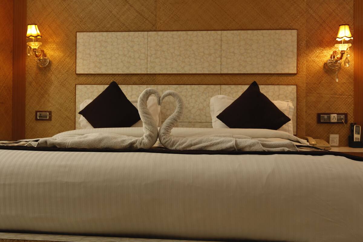 Una habitación de hotel con una cama grande, dos almohadas negras, una disposición de toallas con forma de corazón y lámparas fijadas a la pared.