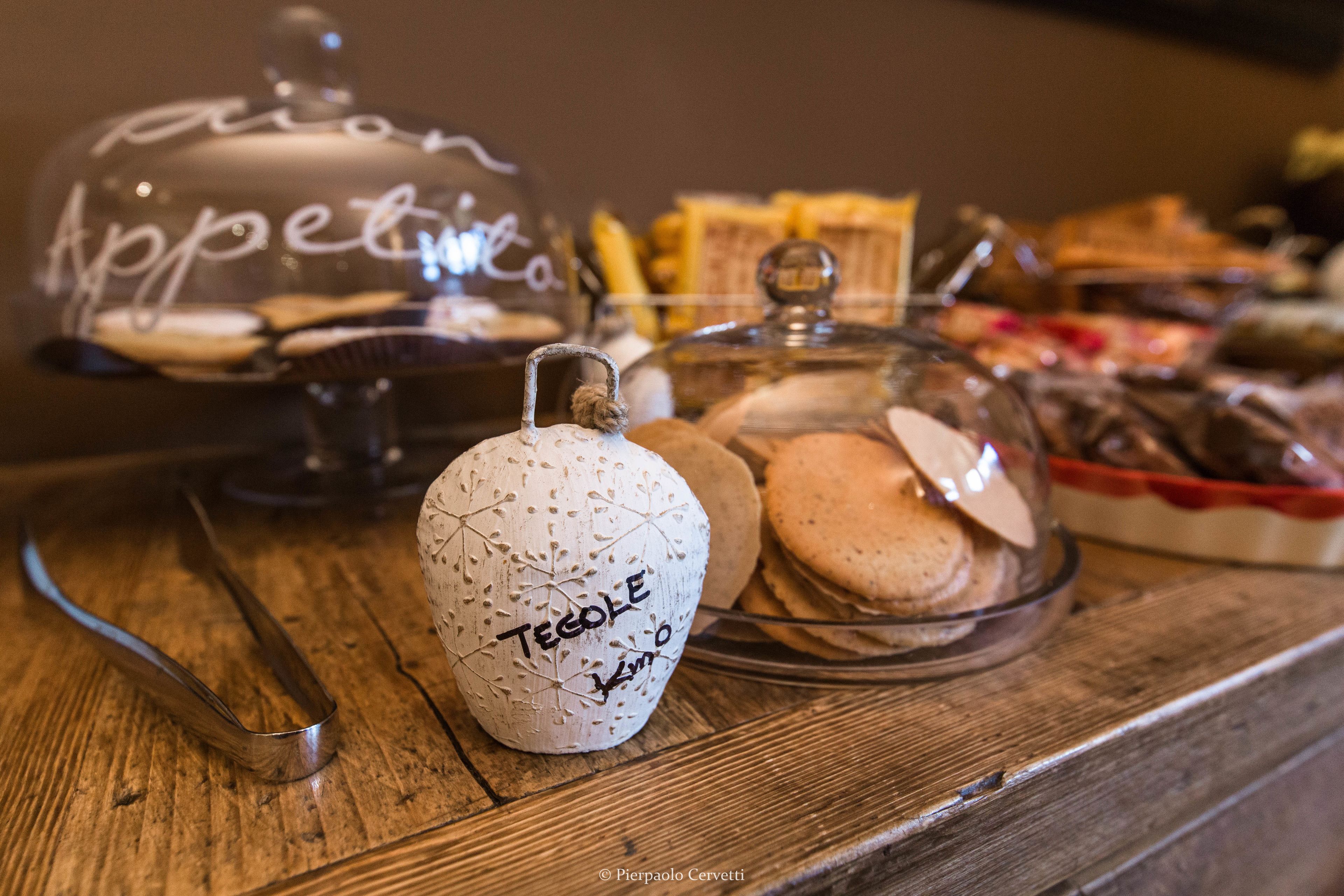 Ein Buffet mit verschiedenen Speisen, darunter eine Glaskuppel mit der Aufschrift "Appetito", ein Keramikbehälter mit "Tegole" und eine Glasschale mit Keksen.