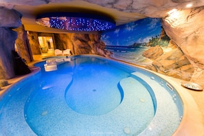 Una amplia piscina interior con un mural de playa tropical, una zona de estar con sillas y un panel de control para las características de la piscina.