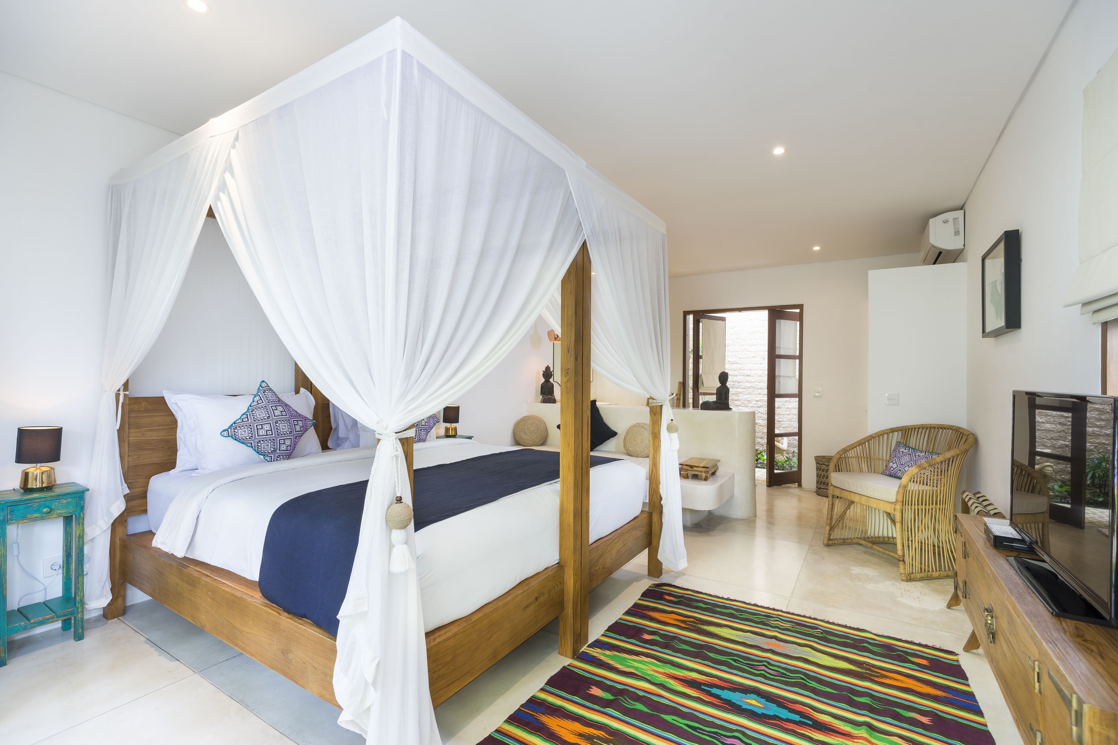 A spacious bedroom with a four-poster bed, a television, a wicker chair, and a colorful rug.