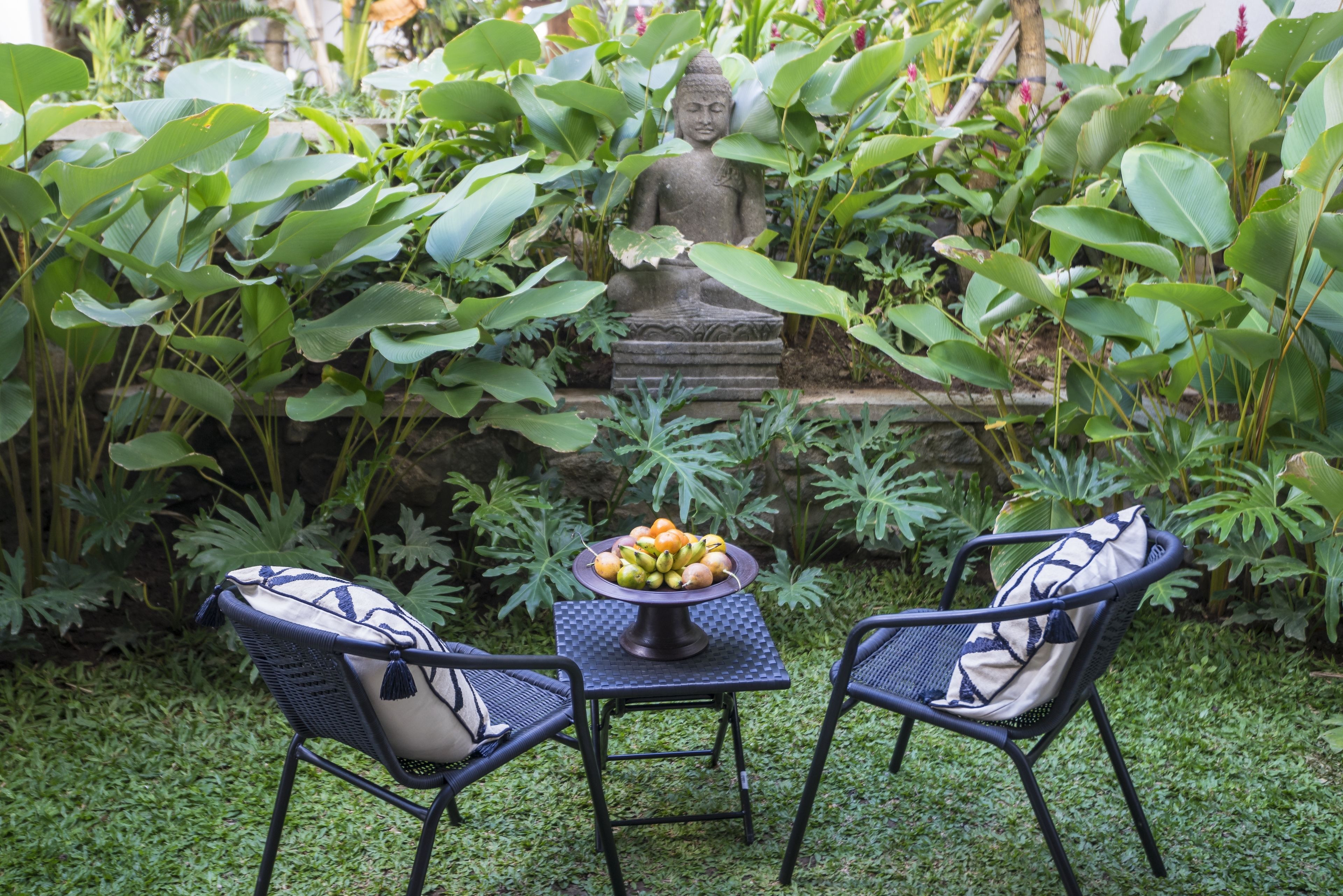 A garden with a Buddha statue, two chairs, and a table with fruit.