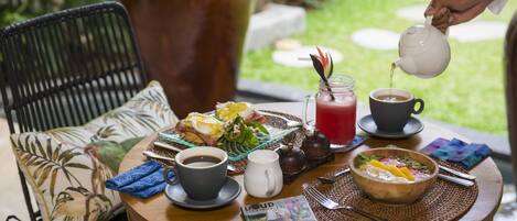 A round wooden table with breakfast items, two cups of coffee, a teapot pouring tea, and a bowl of fruit salad.