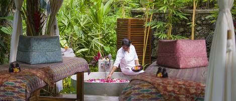A spa area with a woman preparing a bath, surrounded by lush greenery and wooden furniture.