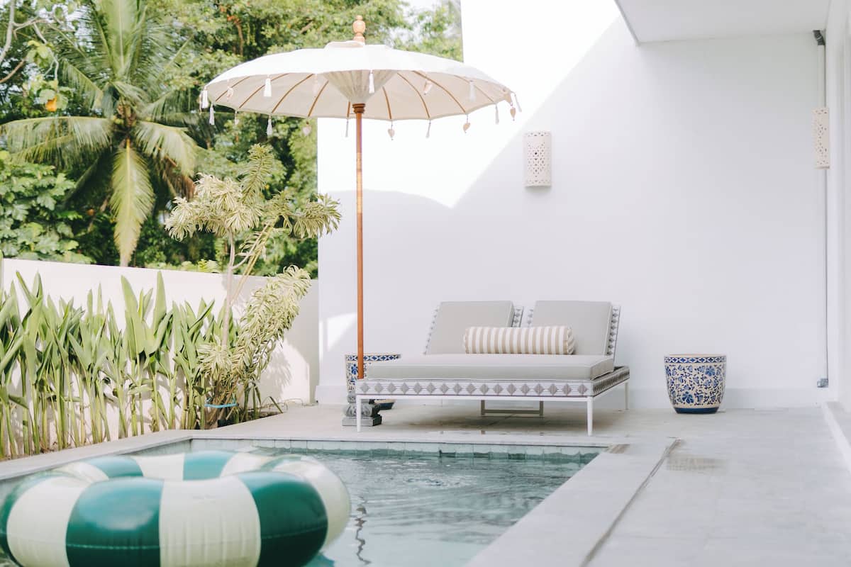 A modern outdoor pool area with a lounge chair and umbrella, surrounded by greenery and a white wall.