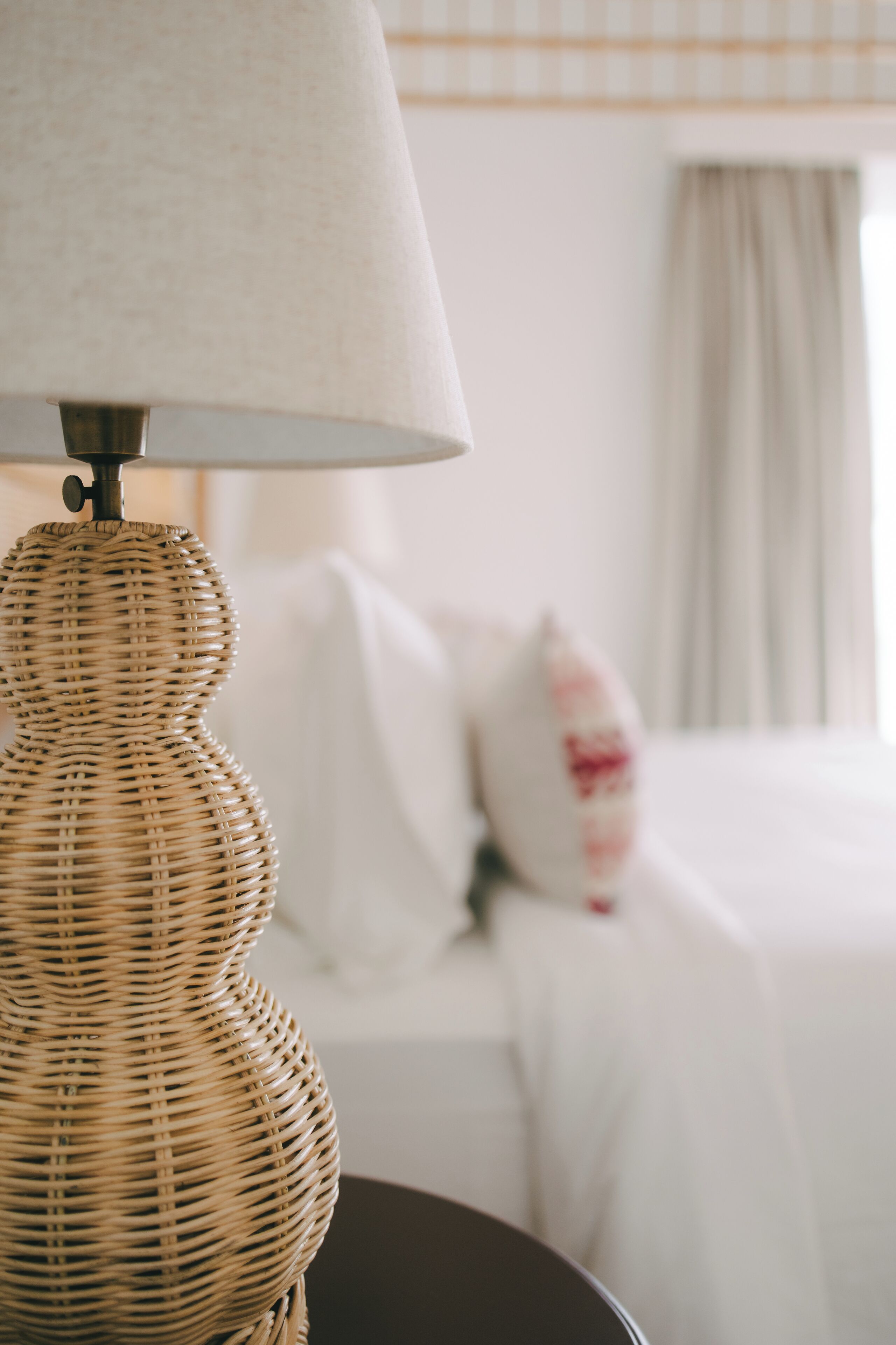 A wicker lamp on a table with a bed in the background.