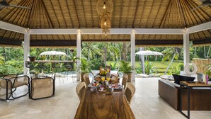A spacious dining area with a wooden table set for breakfast, surrounded by wicker chairs, with a view of lush greenery and a thatched roof overhead. - Calma Ubud (Ubud)