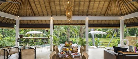 A spacious dining area with a wooden table set for breakfast, surrounded by wicker chairs, with a view of lush greenery and a thatched roof overhead.