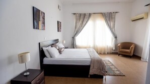 A bedroom with a bed, a chair, a bedside table, and a lamp. - The Gilgal Guest House (Lekki)