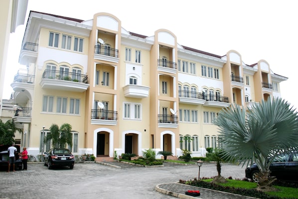 A multi-story residential building with balconies, a paved driveway, and surrounding greenery.