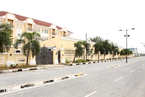 A residential street with a yellow building, a gated entrance, and palm trees.