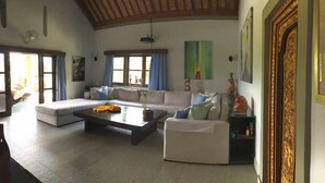 A spacious living room with a large sofa, a coffee table, and a dining area. There is a large window with blue curtains and a ceiling fan. - Villa Sidemen (Sidemen)