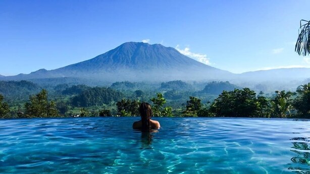 A person in a clear blue infinity pool with a mountain and forest in the background. - Villa Sidemen (Sidemen)