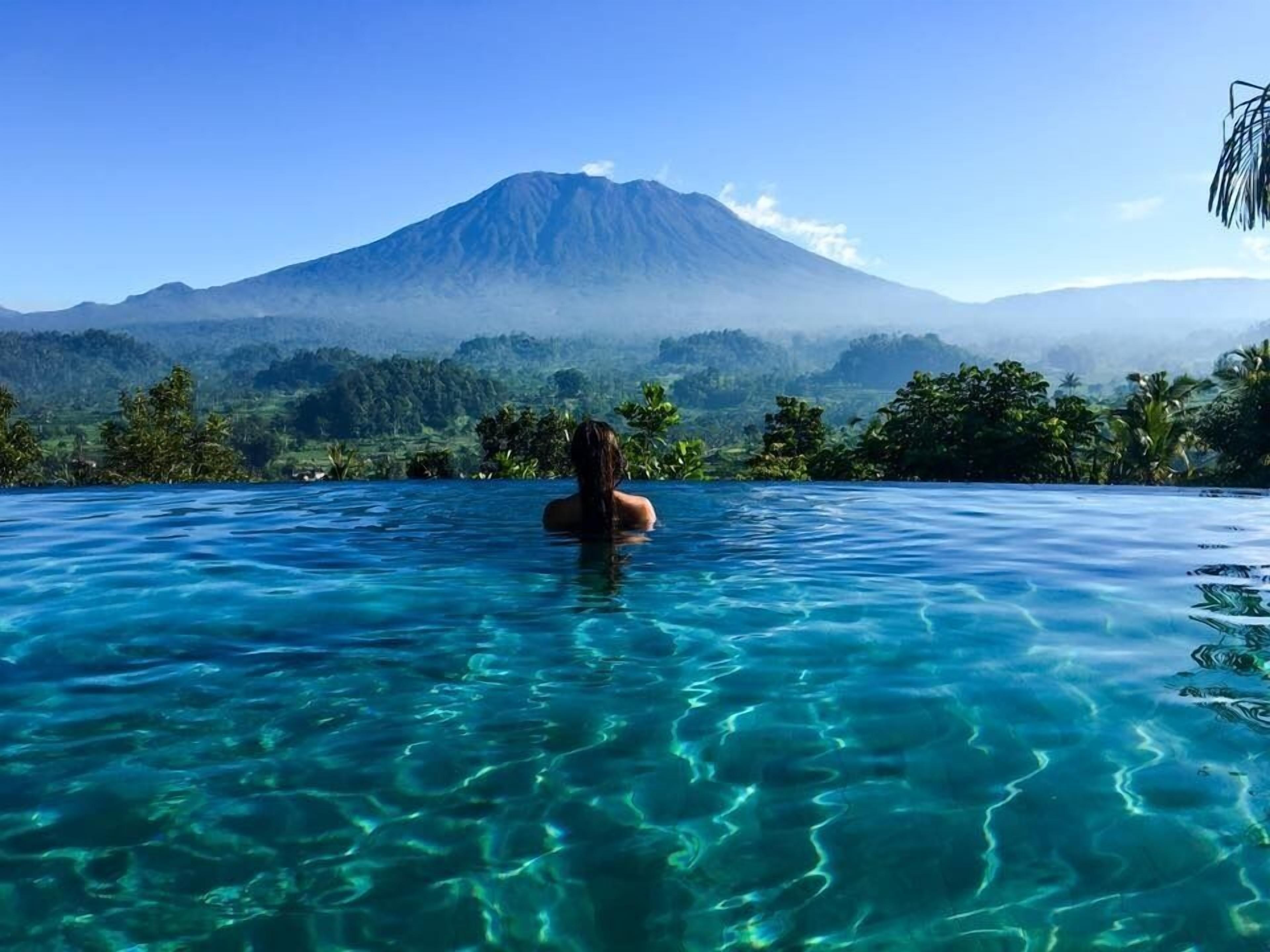 Una persona en una piscina infinita azul cristalina, con una montaña y un bosque al fondo.