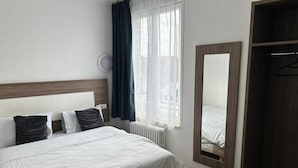 A hotel room with a bed, a table, a chair, a wardrobe, a mirror, and a window with curtains.