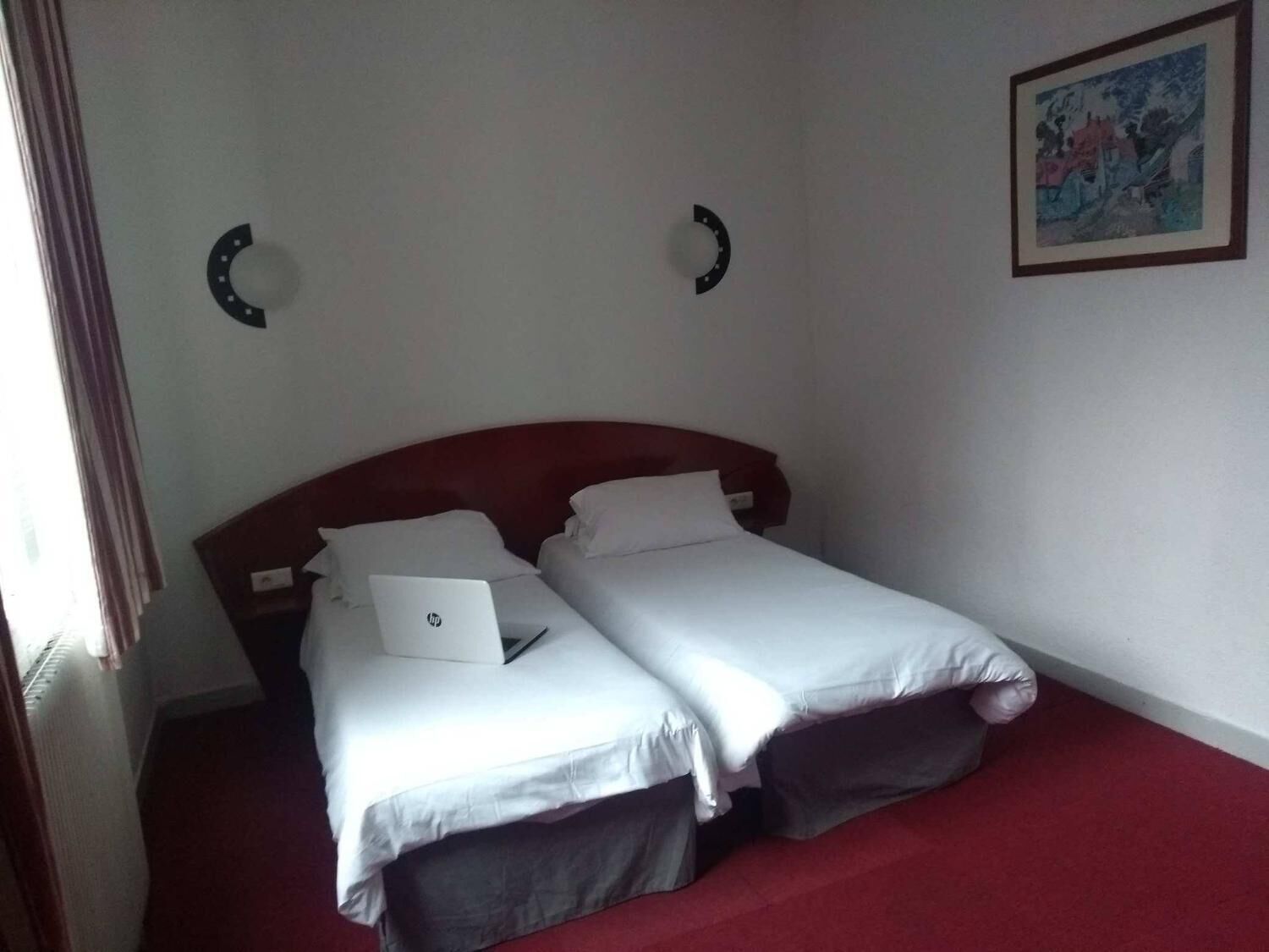 Una cama doble con un ordenador portátil encima, una alfombra roja y un cuadro en la pared.