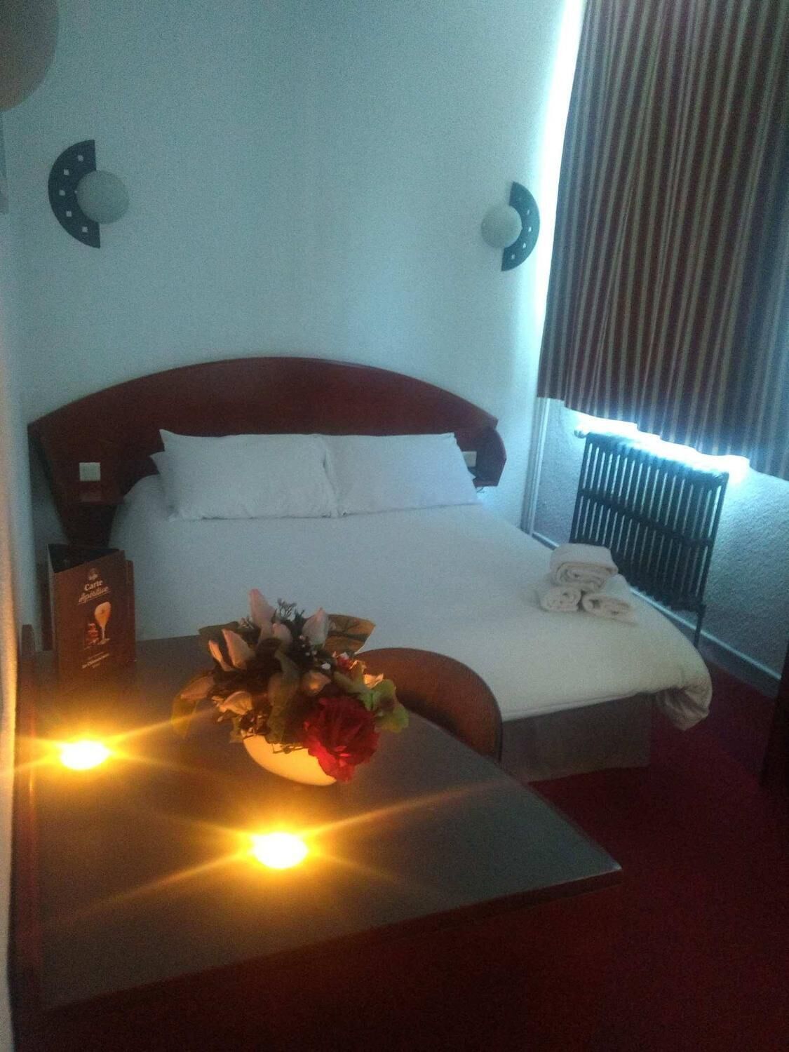 Quarto de hotel com cama, mesa de cabeceira com abajur, arranjo de flores e janela com cortinas.