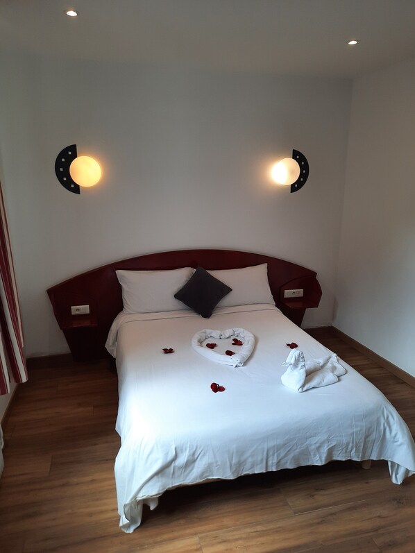A neatly made bed with white linens, a heart-shaped towel, and scattered rose petals. - Hôtel le Clémenceau (Valenciennes)