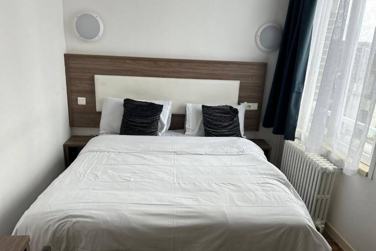 Un lit bien fait, avec une couette blanche, deux oreillers, une tête de lit en bois, des lampes de chevet, une fenêtre avec des rideaux légers et un radiateur.